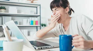 Woman in front of laptop yawning and holding mug